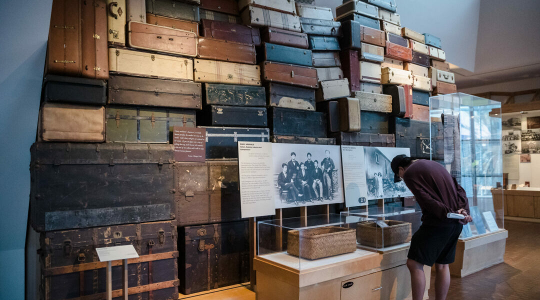 A visitor leans over a display case to look at objects in an exhibition gallery. Behind the case is a wall of suitcases stacked on top of each other.