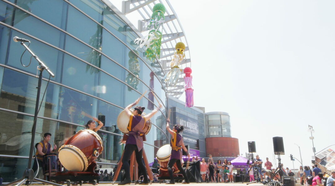 A taiko group performs outside of the Japanese American National Museum. The museum building is behind them in the background with colorful streamers hanging from the top of the building. A large crowd is surrounding the taiko performers.