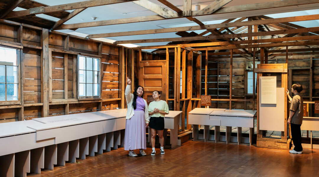 Photo of two people standing in the center of a wooden barrack display. They are looking towards the ceiling and one of the people is pointing up.
