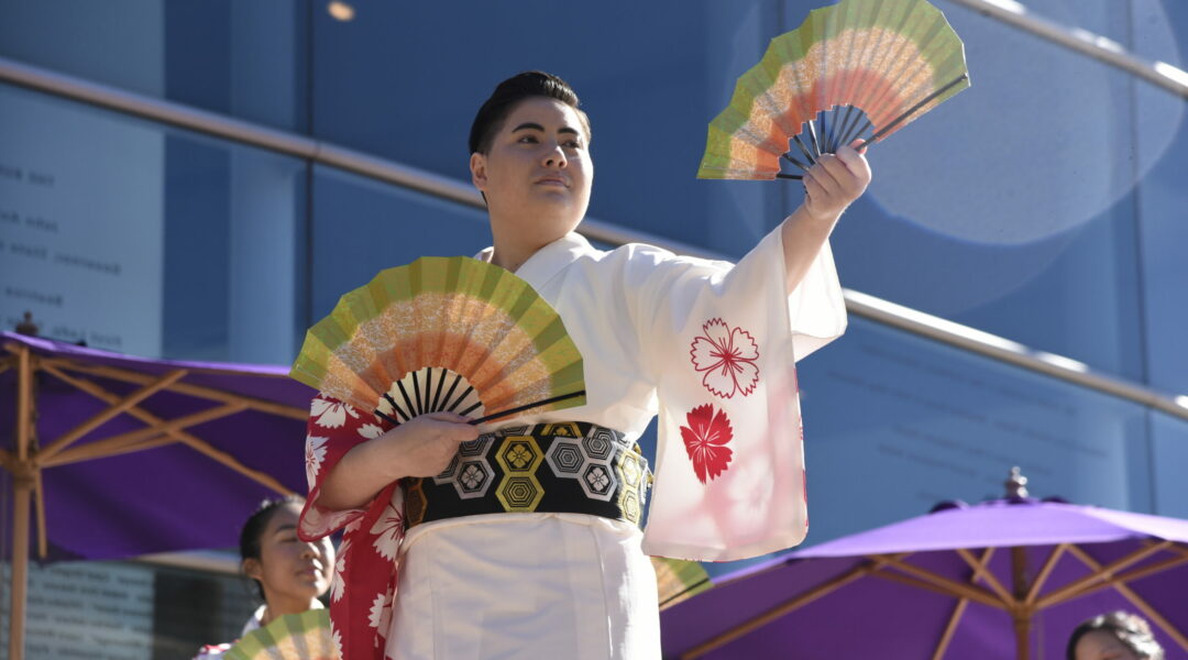 A person wearing a kimono performs a Japanese folk dance while holding Japanese fans in each hand.